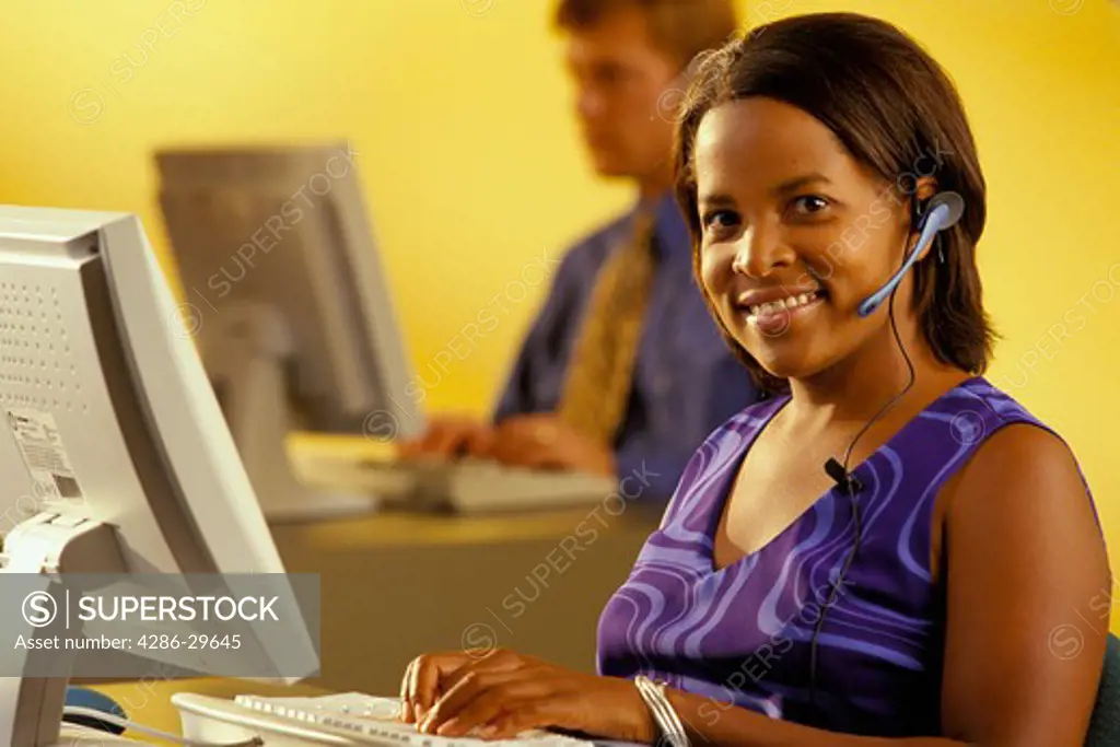 African-American woman wearing a head set while working on her computer and her  male coworker can be seen in the background. 