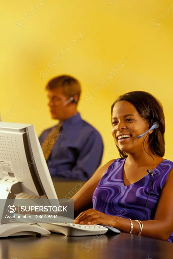 African-American woman wearing a head set while working on her computer and her  male coworker can be seen in the background. 