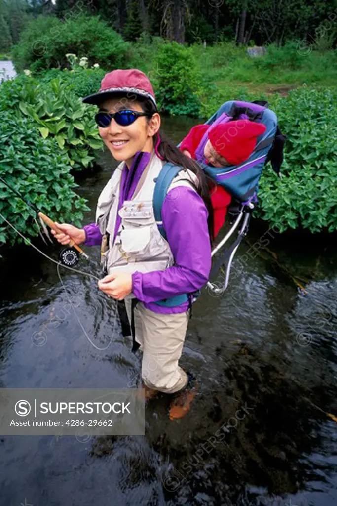 Portrait of a Japanese-American woman fly fishing with her 5 month old daughter on her back in the Metolius River, Oregon. 