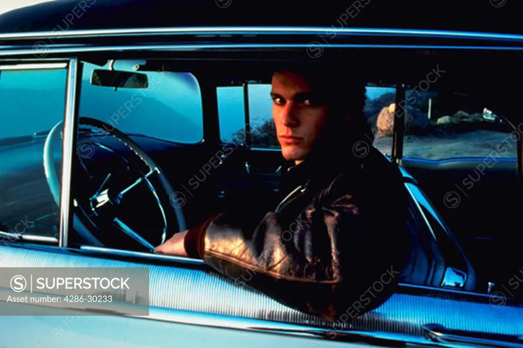 Rebellious-looking young man wearing a leather jacket and sitting in the drivers seat of a classic Cadillac.