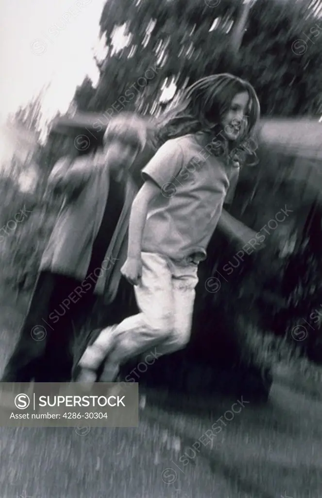Blurred, black and white image of a young girl jumping rope with a young boy in the background swing the jump rope.