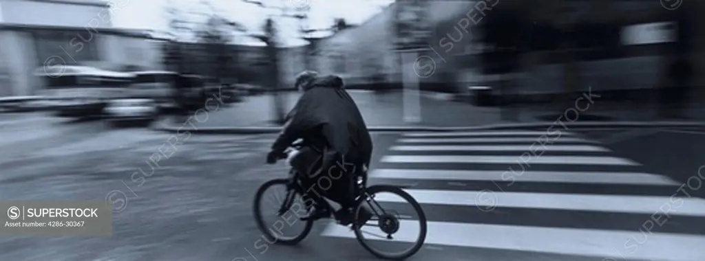 Blurred black and white image of a man riding a bicycle while wearing a rain poncho on a street in Paris, France.