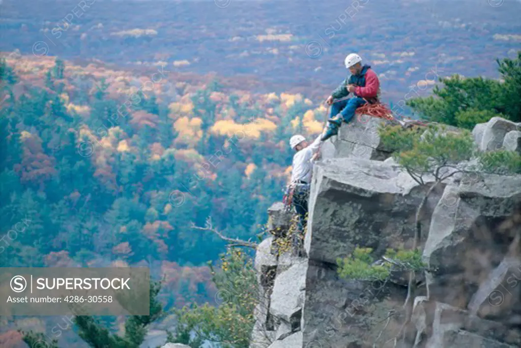 Rockclimber sits on a rock ledge assisting his companion up the cliff face. Behind them a forest shows the beginnings of autumn.