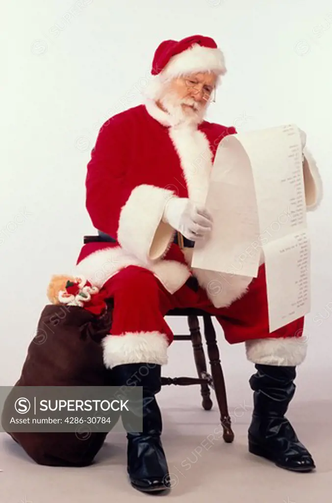 Santa Claus consults a long list of requests as he sits on a stool next to his bag of toys.