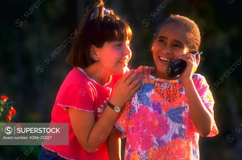 A young girl smiles while talking on a cordless telephone outside as her girlfriend eagerly tries to listen.