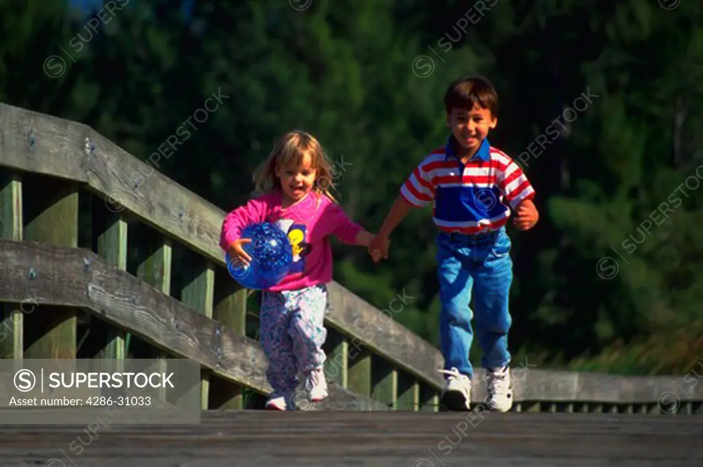 A young boy and girl running on a wooden walkway holding hands. The little girl is holding a ball.