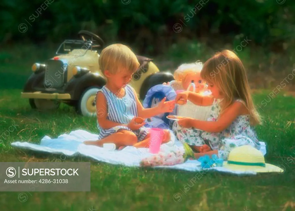 A young boy and girl, age three, sitting on a blanket in the grass having a tea party with a miniature car in the background.