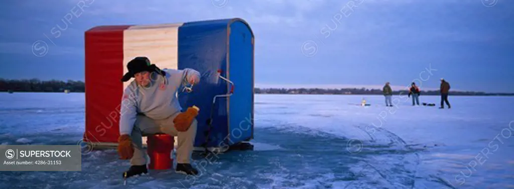 Ice fisherman reeling in line on frozen lake, St. Paul, MN. Red, white and blue ice shack behind him.