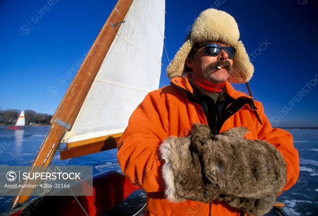 Man sits on his ice sailboat on a frozen lake wearing fur hat and gloves, Minnesota.