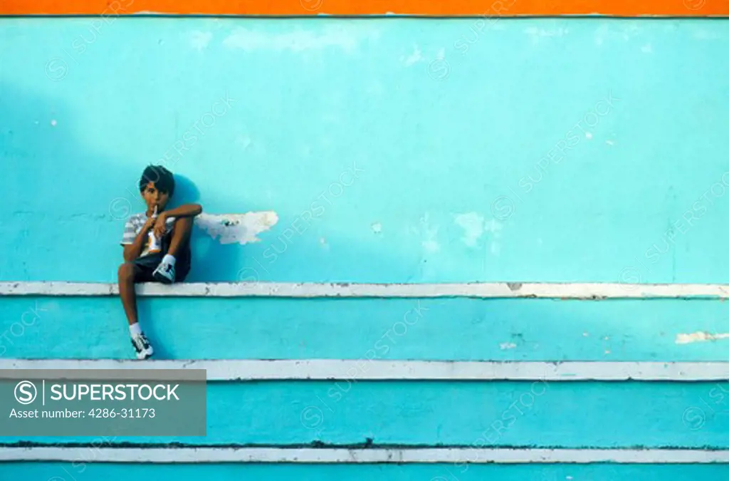 Young Mexican boy sits on painted steps of bleechers drinking soda at a playground, Isla Mujeres, Mexico.