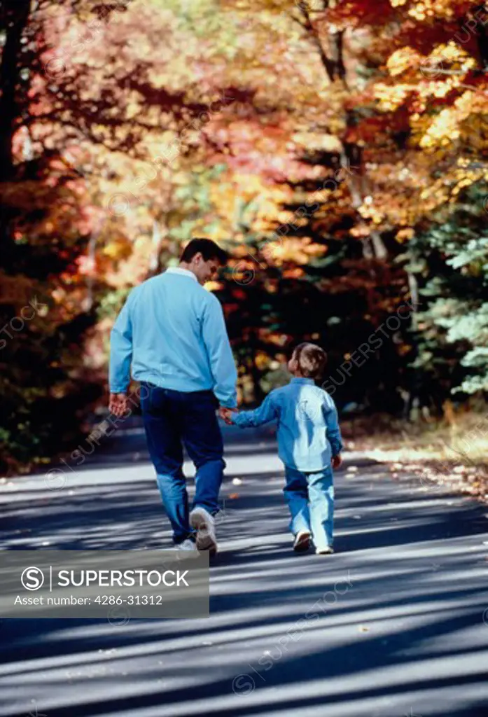 Father and young son holding hands and walking along country road with fall foliage.