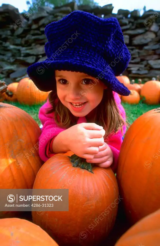Portrait of a young girl wearing a big blue hat and sitting in a pumpkin patch.