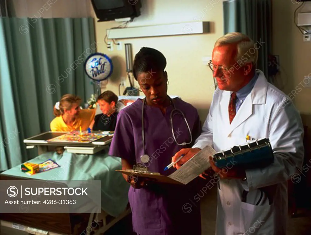A nurse and doctor review a patients chart while a young boy in a hospital bed and his mother play a board game in the background.