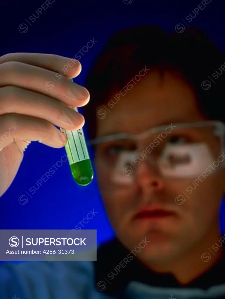 A lab tech wearing protective gloves and protective eye wear holding up a test tube vial containing a green fluid.