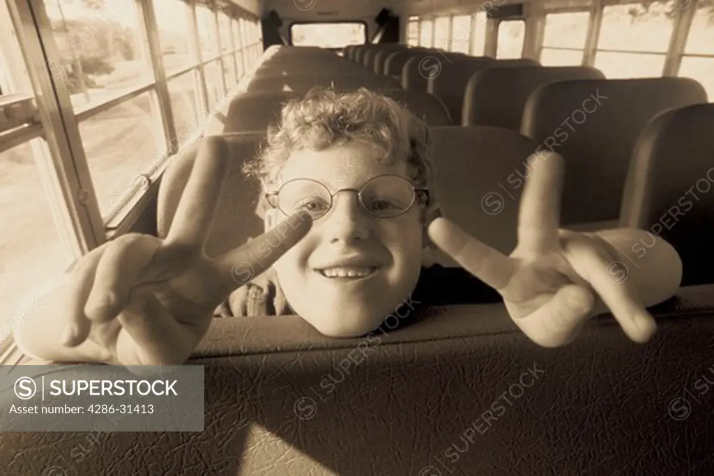 Black and white close-up of a child leaning back on a bus seat giving the peace signs with both hands. 