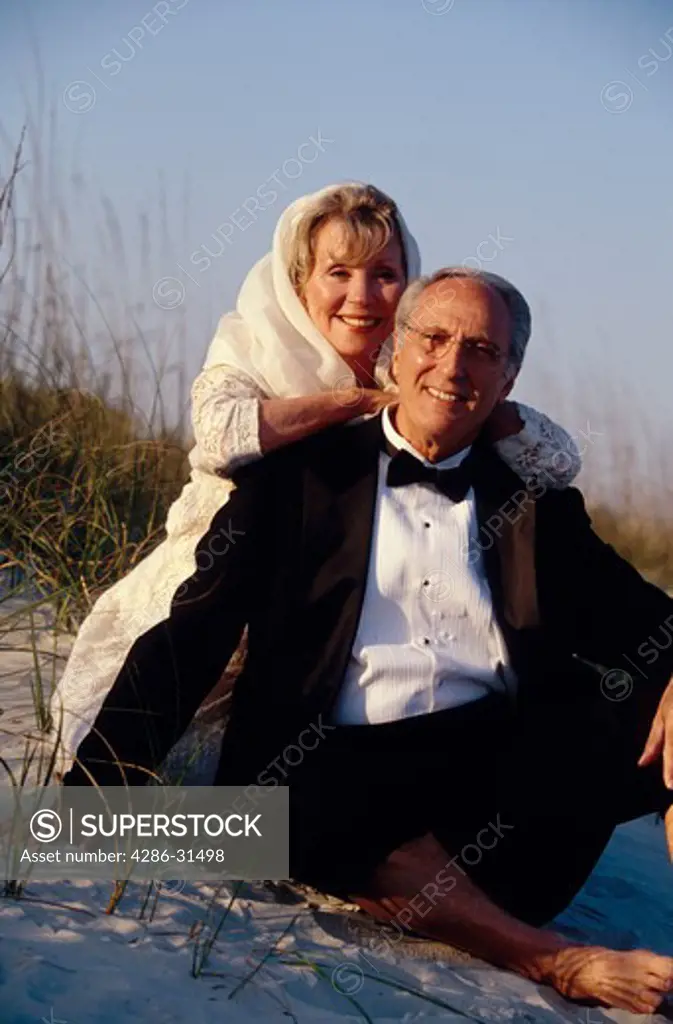 Smiling mature couple wearing a tuxedo and formal gown sitting together on a sand dune on a beach.