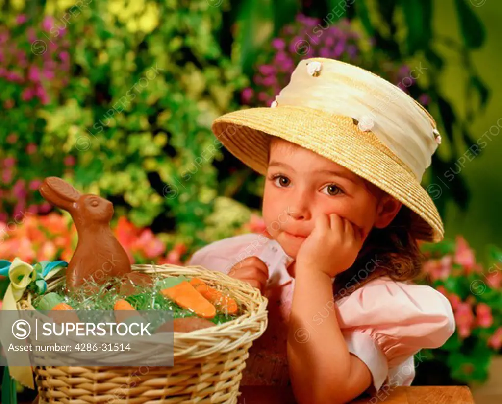 Portrait of a little girl wearing her Easter bonnet sitting beside a basket containing a chocolate bunny and other candies.