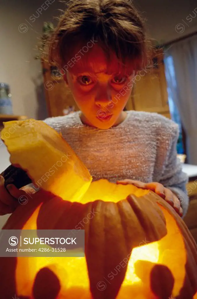 A girl lifts the lid of a carved jack-o'-lantern and her face is lit from below by the candlelight.