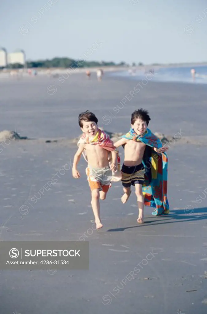 Young twin brothers wear capes made from beach towels as they run together along a beach.
