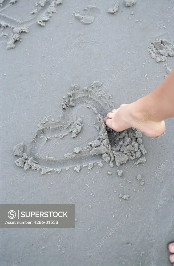 A person uses their toes to draw a heart shape in the wet sand at a beach.
