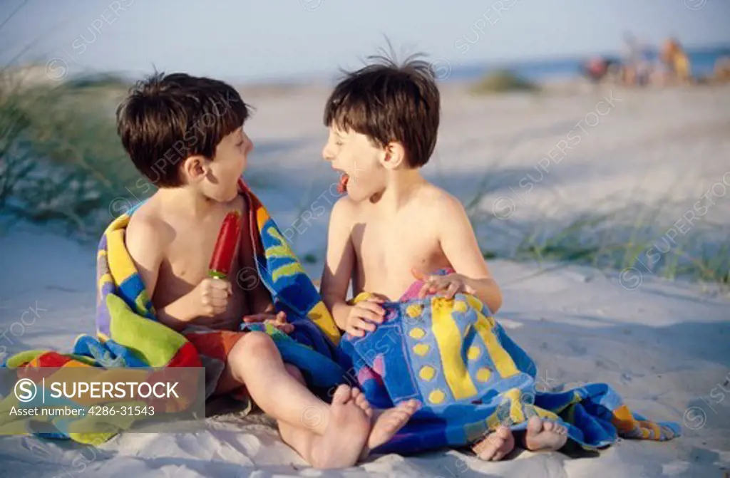 Twin brothers stick their tongues out at each other while enjoying popsicles at a beach.