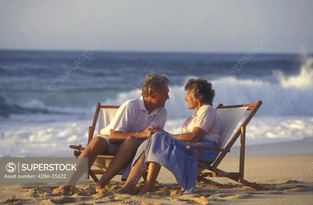 Mature couple holding hands and gazing into each others eyes as they sit in folding chairs on the beach.