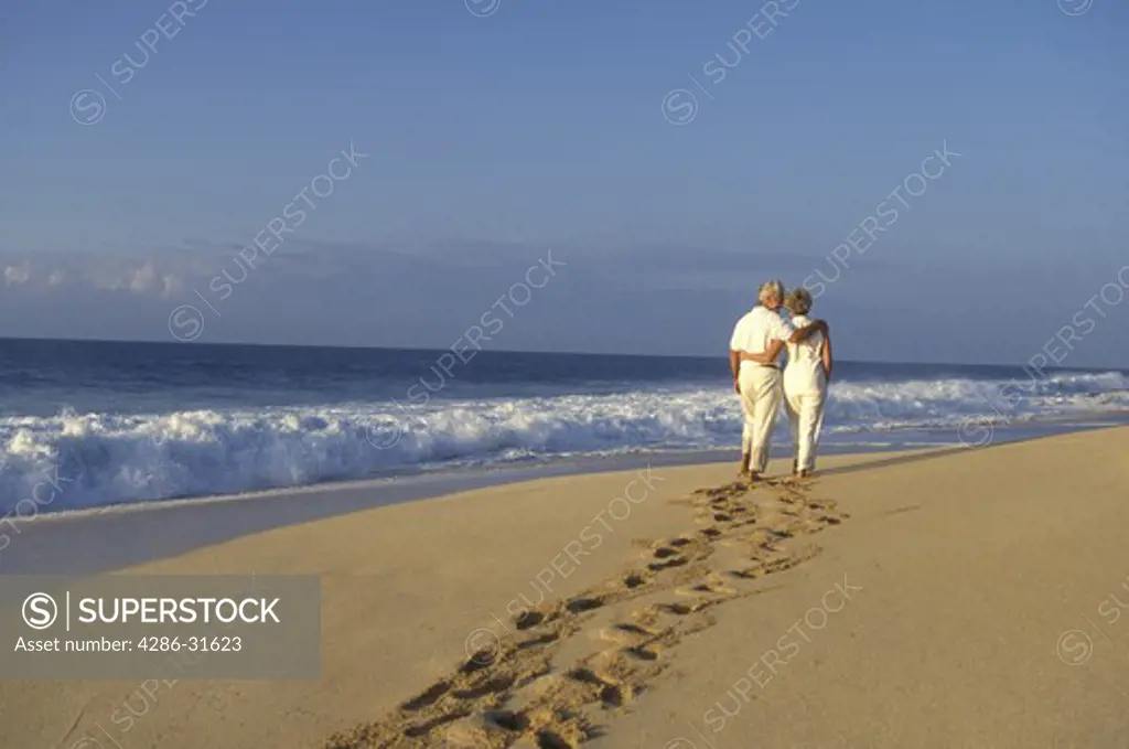 Mature couple leave deep footprints in the sand as they walk together down an empty beach.