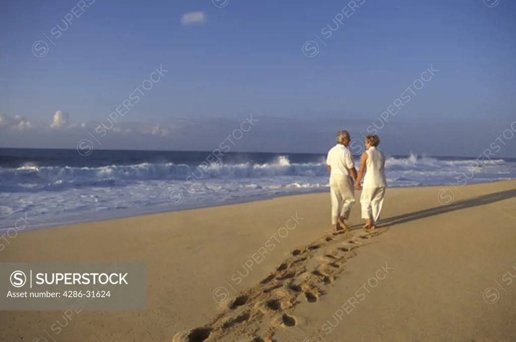 Mature couple leave deep footprints in the sand as they walk together down an empty beach while holding hands.