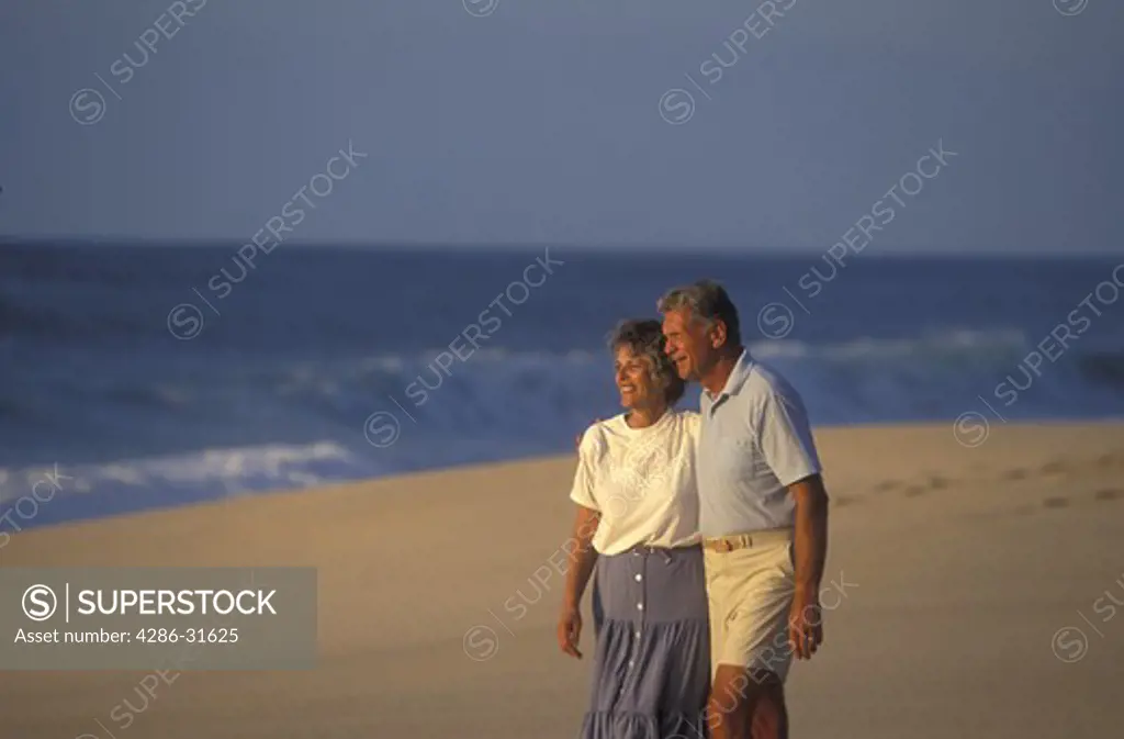 Casually dressed mature couple look towards the sun as they walk together on a wide sandy beach.