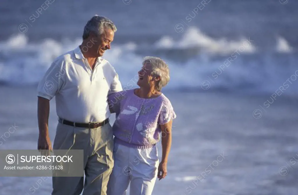 Casually dressed mature couple smile at each other as they walk together along the beach as surf crashes in the background.
