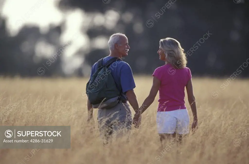Casually dressed mature couple smile at each other as they walk hand-in-hand through hip-high yellow grasses.