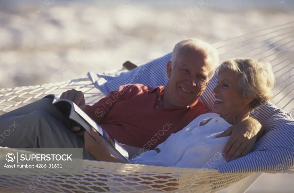 Retired couple happily reading a magazine as they lie together in a hammock outdoors on a sunny day.