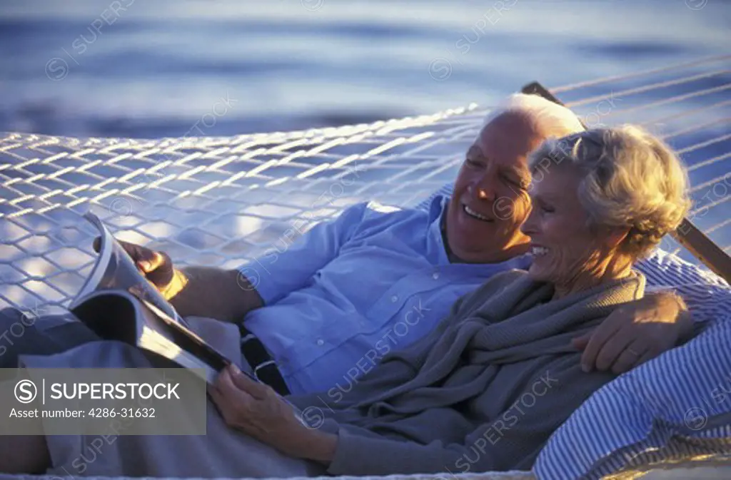 Retired couple happily reading a magazine as they lie together in a hammock outdoors on a sunny day.