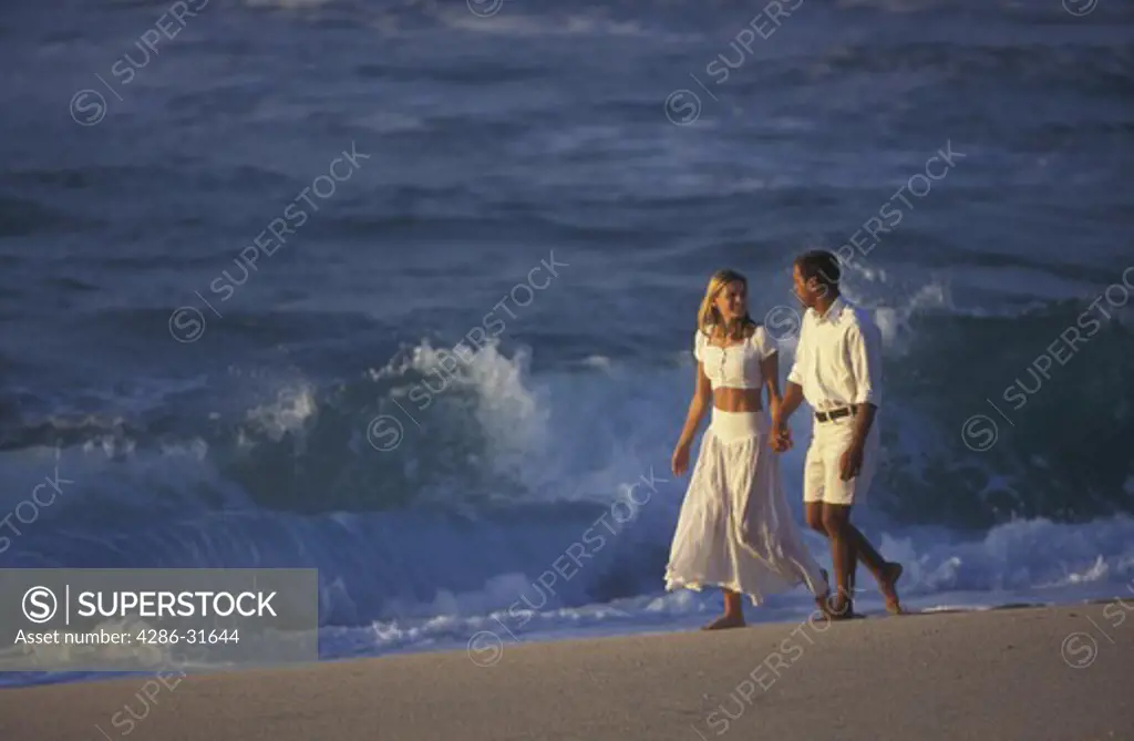 Casually dressed barefoot couple look at each other as they walk hand-in-hand on a beach as waves break in the background.