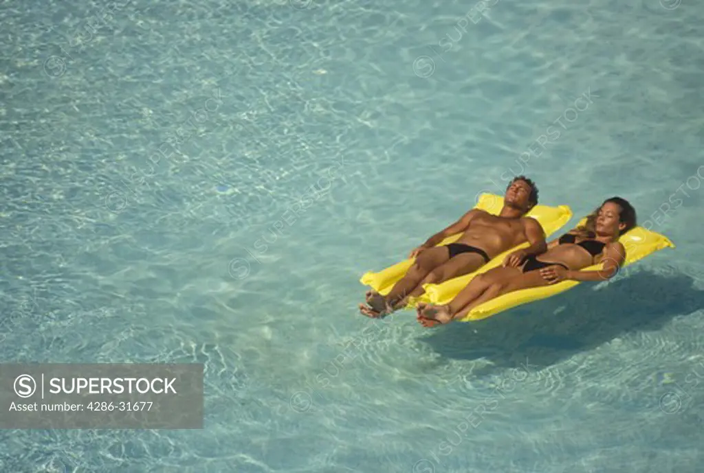 Couple floating side-by-side on yellow inflatable rafts in a large swimming pool.