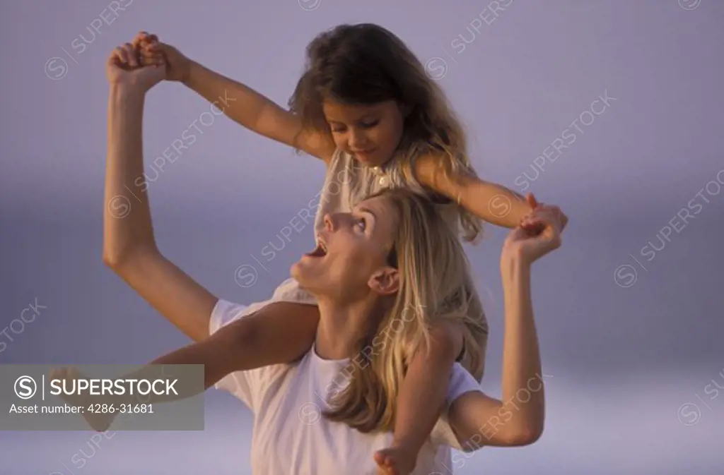 Woman looks up at the young daughter she is carrying outdoors on her shoulders.