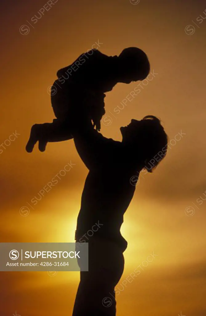 A mother holds her young child aloft over her head while being silhouetted by yellow sunlight.