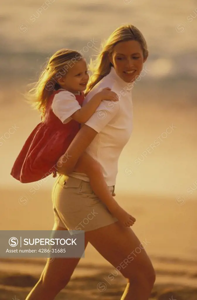 A mother carries her young daughter piggyback as they walk along the beach.