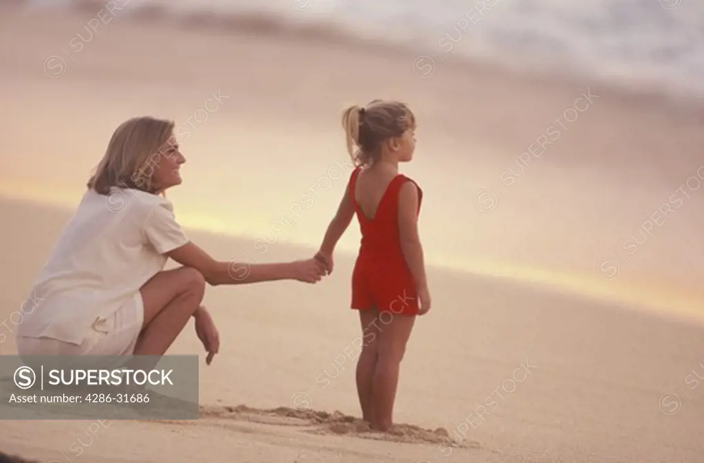 A mother holds her young daughters hand as they look out from the beach to the ocean beyond.