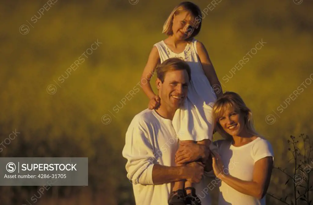 Outdoor family portrait of a mother and father standing together as he lifts his daughter up onto his shoulder.