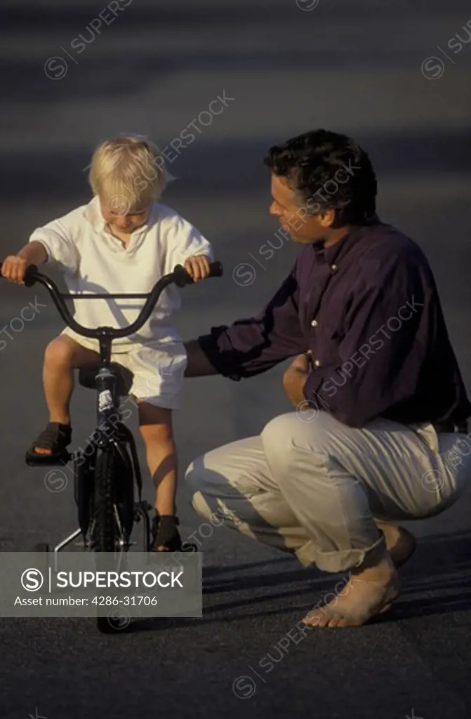Young boy concentrates on balancing his bicycle with training wheels as his barefoot father gives words of encouragement.