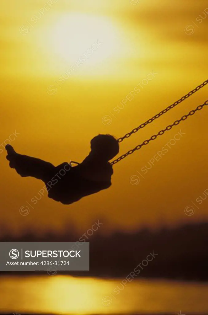 Silhouetted by the setting sun, a young child soars high on a swing.