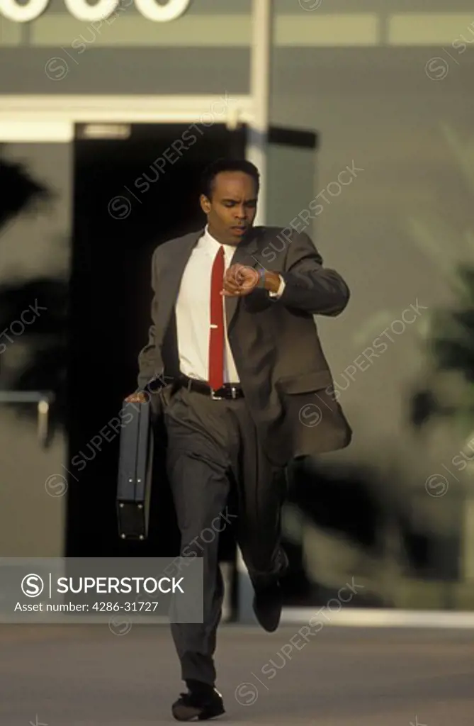 African-American businessman looks at his watch as he runs briefcase in hand out of an office building.