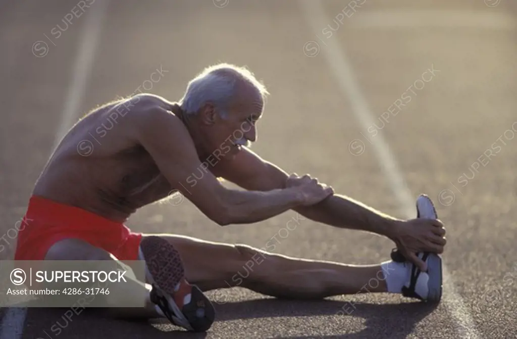 Senior shirtless man doing stretching sit-ups on an outdoor track.