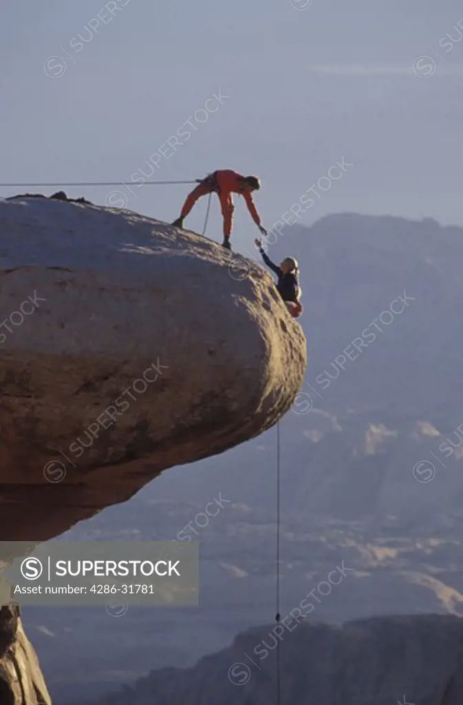 Male climber on a safety rope reaches out to a female climber as she makes her way up and over an outcrop of rock at the summit.
