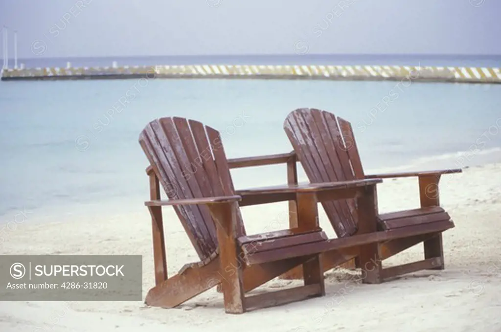 Two adirondack chairs sit on a beach in Cozumel, Mexico by the calm water.