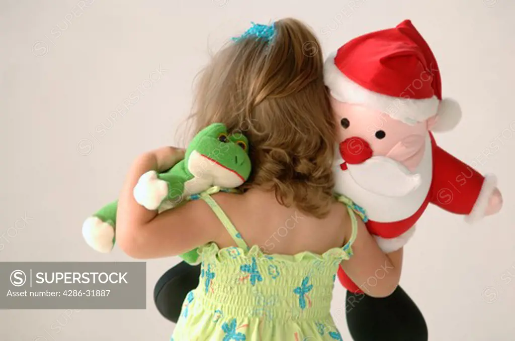 Back of a 2 year old girl holding a stuffed toy frog and a stuffed toy Santa Claus. 