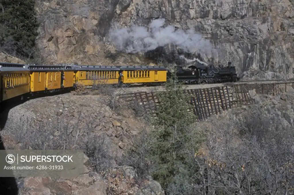 The Durango & Silverton Narrow Gauge Railroad, a historic steam powered locomotive, passes by steep rocky cliffs.