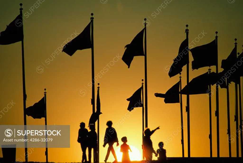Tourists and flags on the grounds of the Washington Monument are silhouetted by the setting yellow sun.