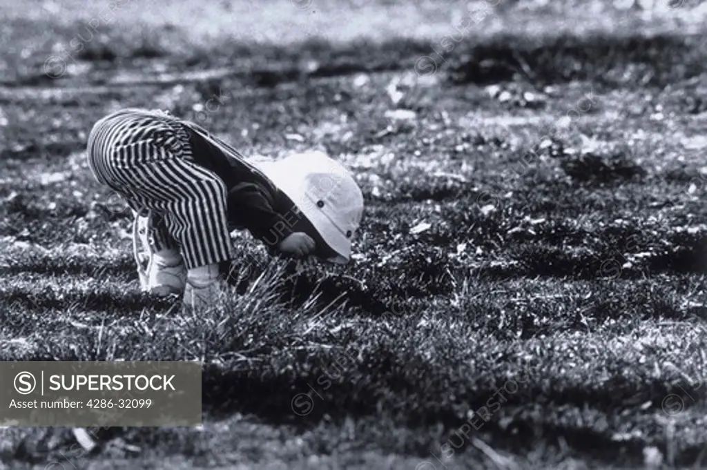 Young 2-year-old girl wearing a beach hat and bending down to smell a flower in the grass.